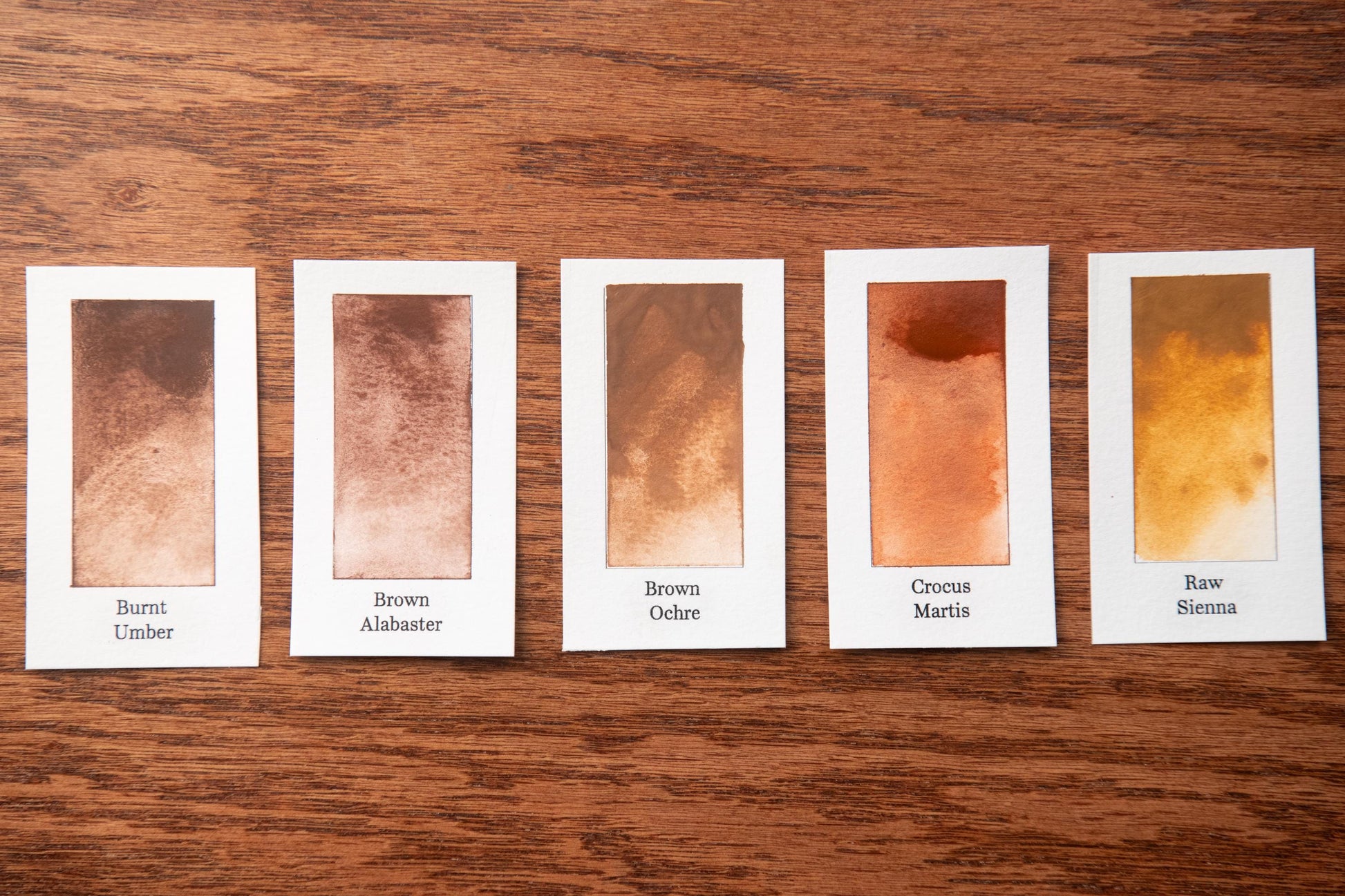 five small rectangular cards with different shades of brown paint, arranged on a wooden surface.