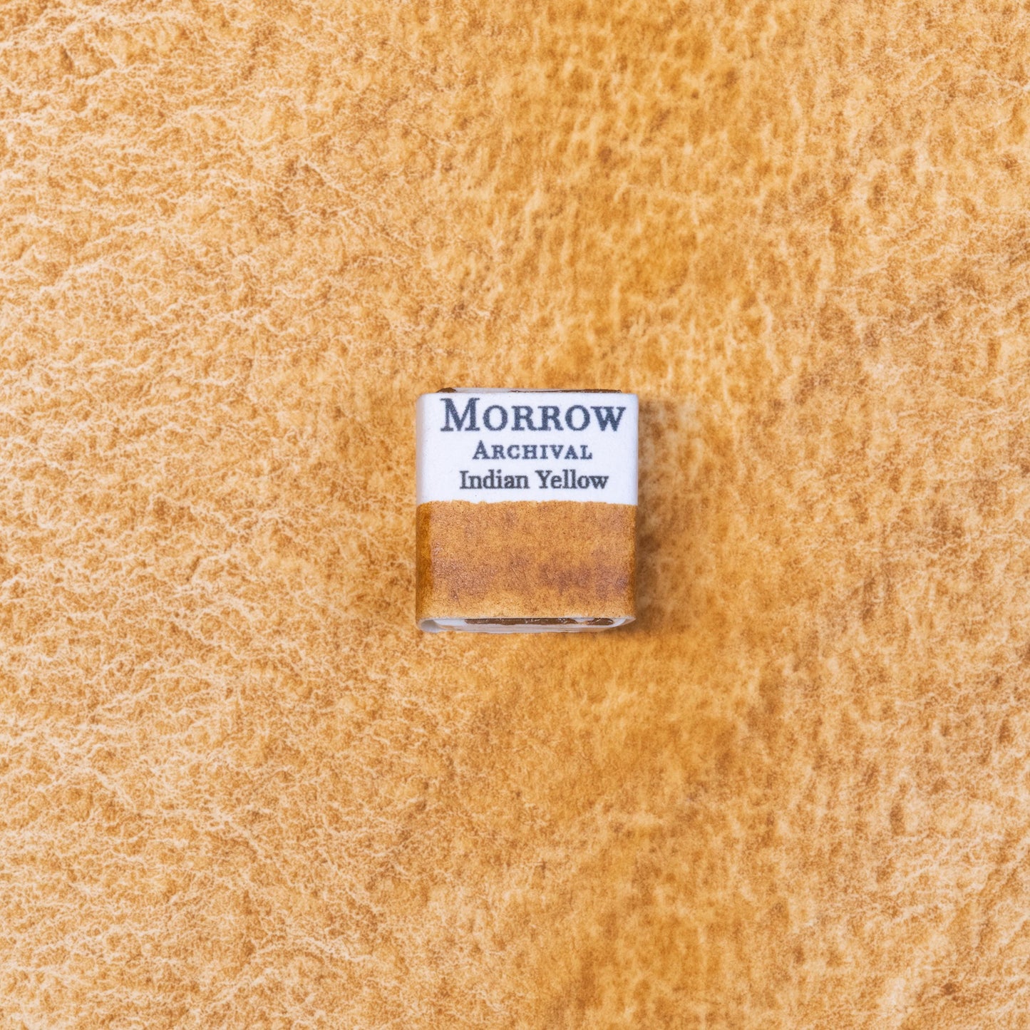 Close-up image of a half pan of Indian Yellow handmade watercolor paint, showing a rich, golden-yellow hue with a smooth, luminous surface. The pan sits on a neutral or rustic background, emphasizing its warm glow and handcrafted quality. Ideal for artists seeking a radiant, transparent yellow with historical roots and modern performance.