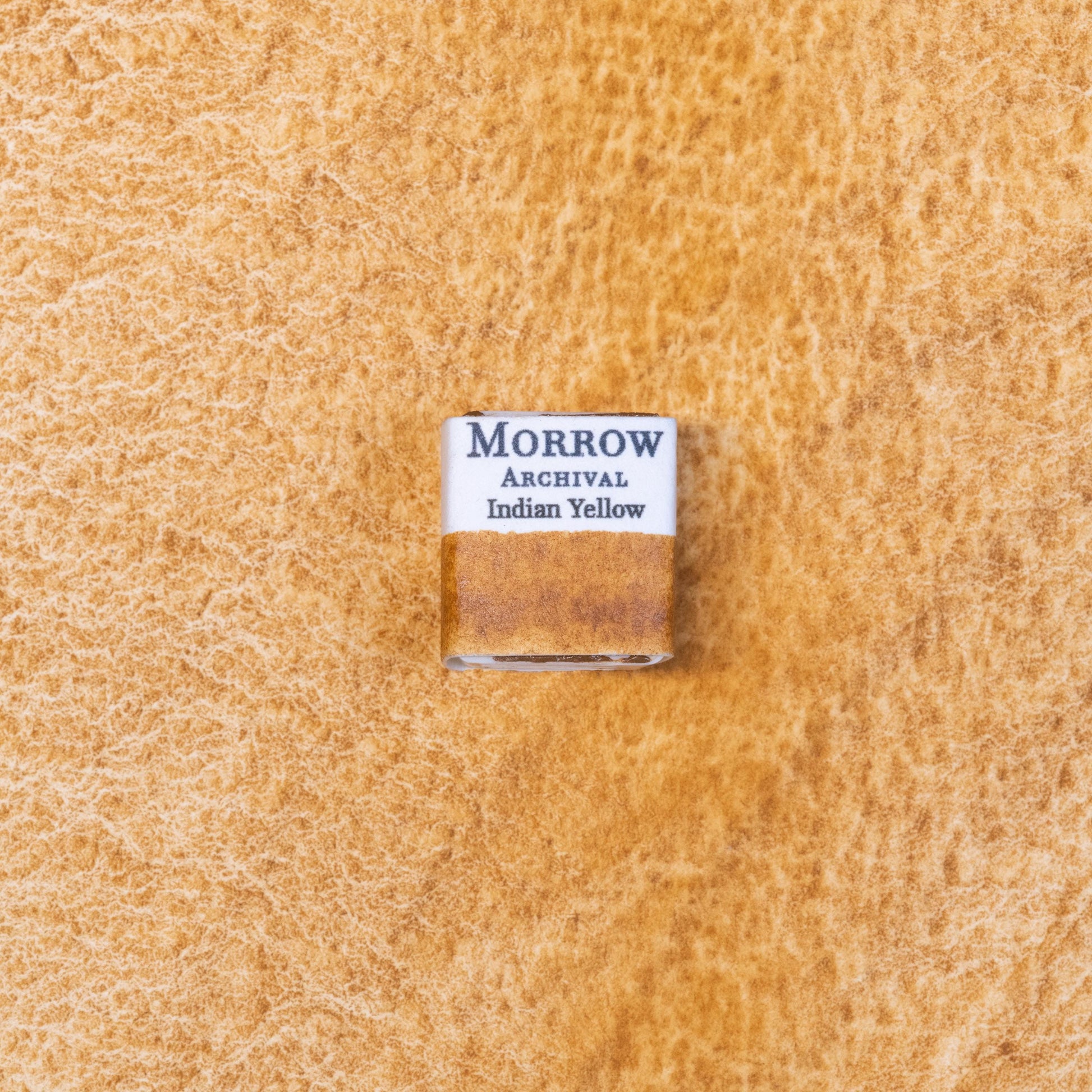 Close-up image of a half pan of Indian Yellow handmade watercolor paint, showing a rich, golden-yellow hue with a smooth, luminous surface. The pan sits on a neutral or rustic background, emphasizing its warm glow and handcrafted quality. Ideal for artists seeking a radiant, transparent yellow with historical roots and modern performance.