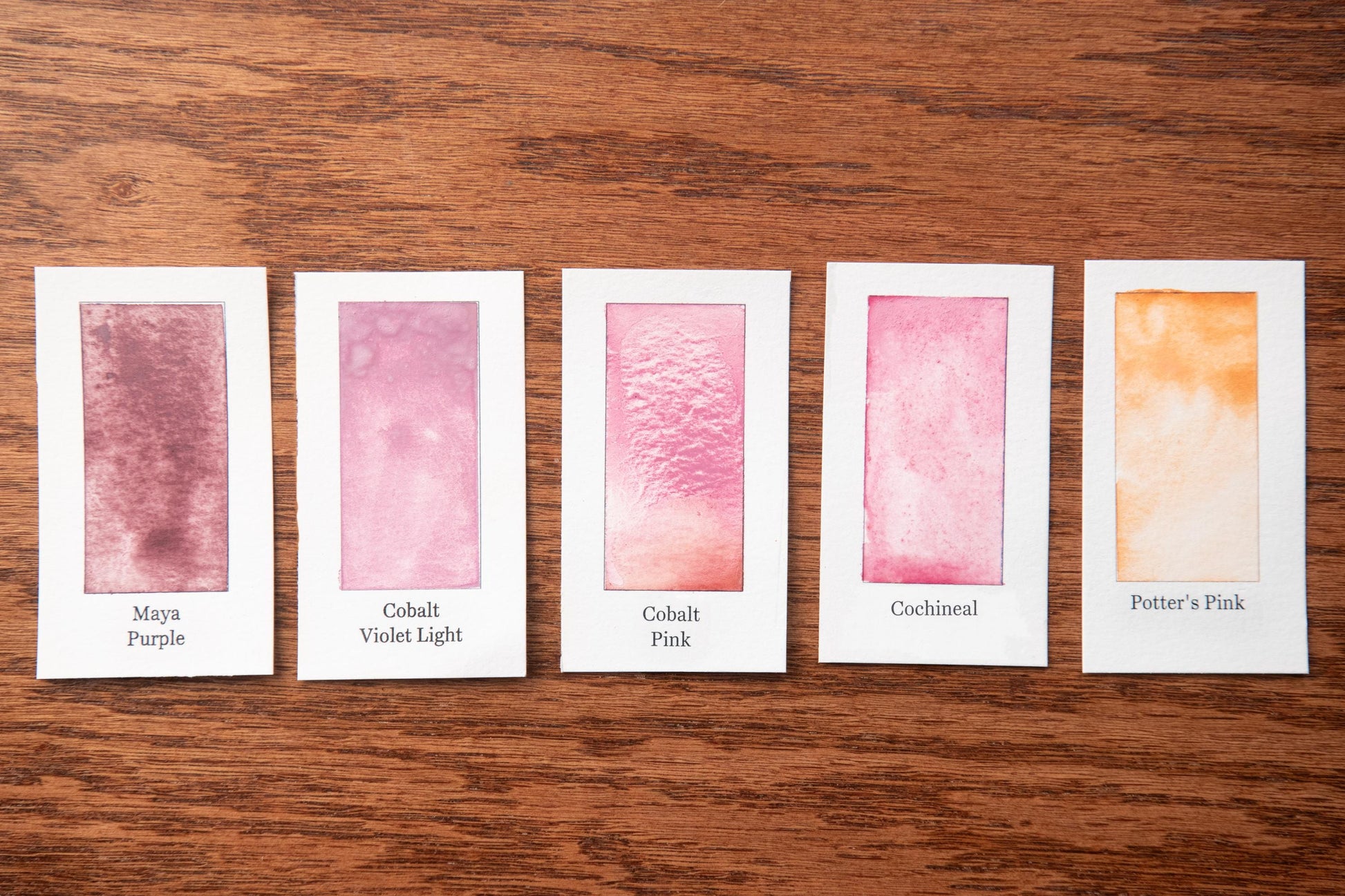 five watercolor paintings with different shades of pink, arranged on a wooden surface.