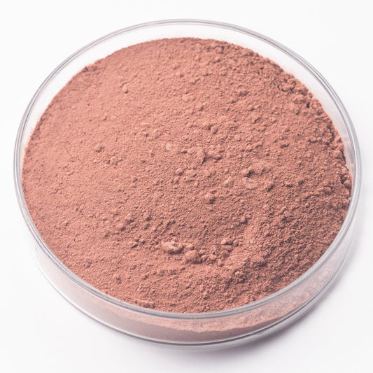 A clear petri dish holds Sorghum Lake Pigment, a handmade, plant-based reddish-brown powder with a smooth texture and some small clumps, displayed on a white background.