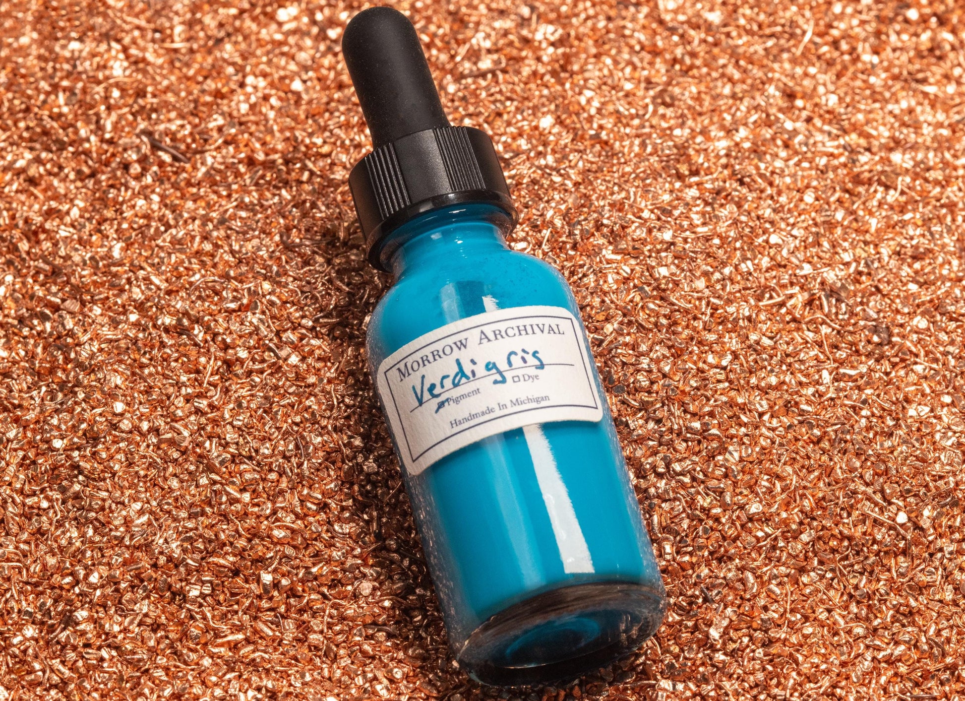 Verdigris ink bottle resting on oxidized copper sheets and fine copper acetate powder, visually tying the vibrant turquoise ink to its chemical-mineral origins. The scene reflects historic methods of pigment creation from patinated metal.