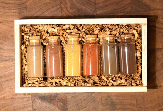 Set of 6 all natural ochre earth pigments in 1oz glass bottles with corks, nestled in shredded recycled kraft paper inside a handcrafted wooden gift box. Resting on a dark walnut wood board with soft, moody lighting, this all-natural artist gift set is ideal for birthdays, holidays, or creative milestones. Each bottle contains rich earth pigments in tones from golden yellow to deep green and warm brown as well as red and orange and are archival quality and lightfast for painting.