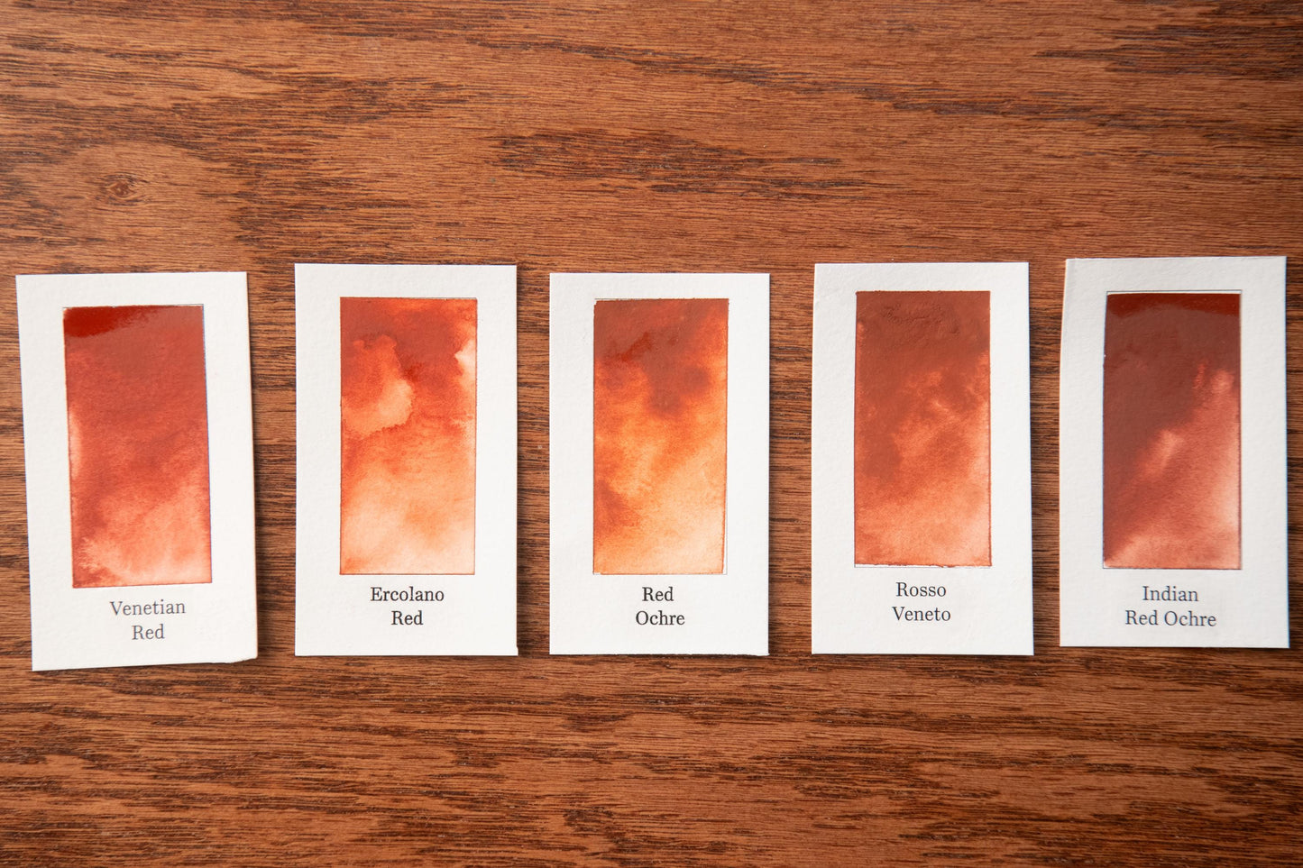 five identical cards, each containing a different shade of red paint. The cards are arranged in a row on a wooden surface.