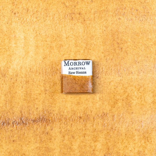 Close-up image of a half pan of Raw Sienna handmade watercolor paint, showing a warm, golden-brown hue with a smooth matte finish and gentle granulation. The pan is set on a neutral background, emphasizing its natural earth origin and artisanal craftsmanship. Ideal for artists seeking a classic, lightfast earth pigment with warmth and versatility.
