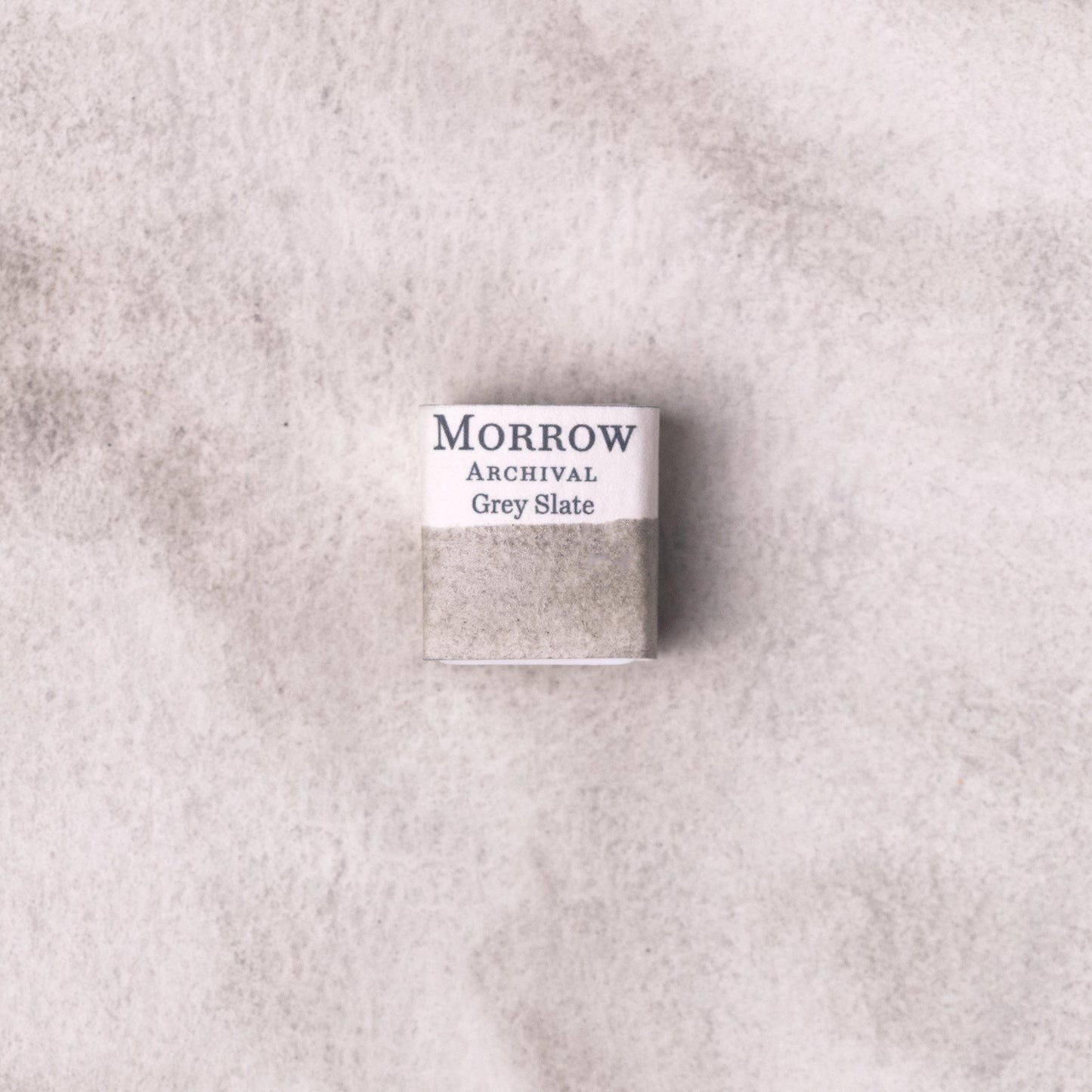 Handmade Slate Grey natural watercolor paint half pan resting on a textured watercolor painting. Crafted from pure single-pigment natural slate stone, this cool, neutral grey offers subtle granulation and excellent transparency, ideal for tonal studies, landscape painting, and historical illustration. A timeless eco-friendly gift for watercolor artists and collectors seeking small-batch, archival-quality, all-natural handmade pigments sourced from real stone.