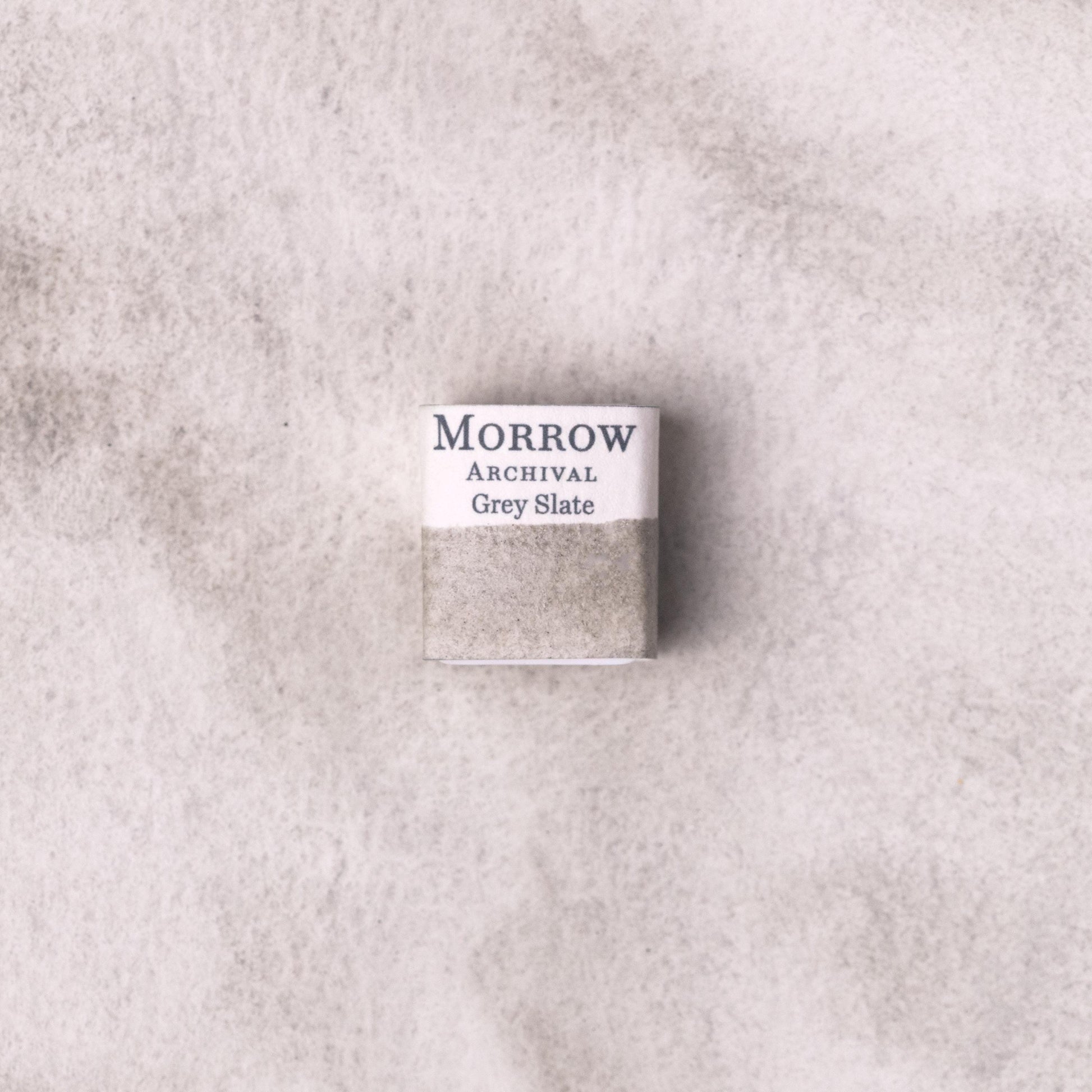 Handmade Slate Grey natural watercolor paint half pan resting on a textured watercolor painting. Crafted from pure single-pigment natural slate stone, this cool, neutral grey offers subtle granulation and excellent transparency, ideal for tonal studies, landscape painting, and historical illustration. A timeless eco-friendly gift for watercolor artists and collectors seeking small-batch, archival-quality, all-natural handmade pigments sourced from real stone.