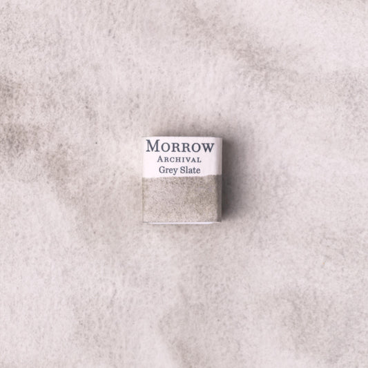 Handmade Slate Grey natural watercolor paint half pan resting on a textured watercolor painting. Crafted from pure single-pigment natural slate stone, this cool, neutral grey offers subtle granulation and excellent transparency, ideal for tonal studies, landscape painting, and historical illustration. A timeless eco-friendly gift for watercolor artists and collectors seeking small-batch, archival-quality, all-natural handmade pigments sourced from real stone.