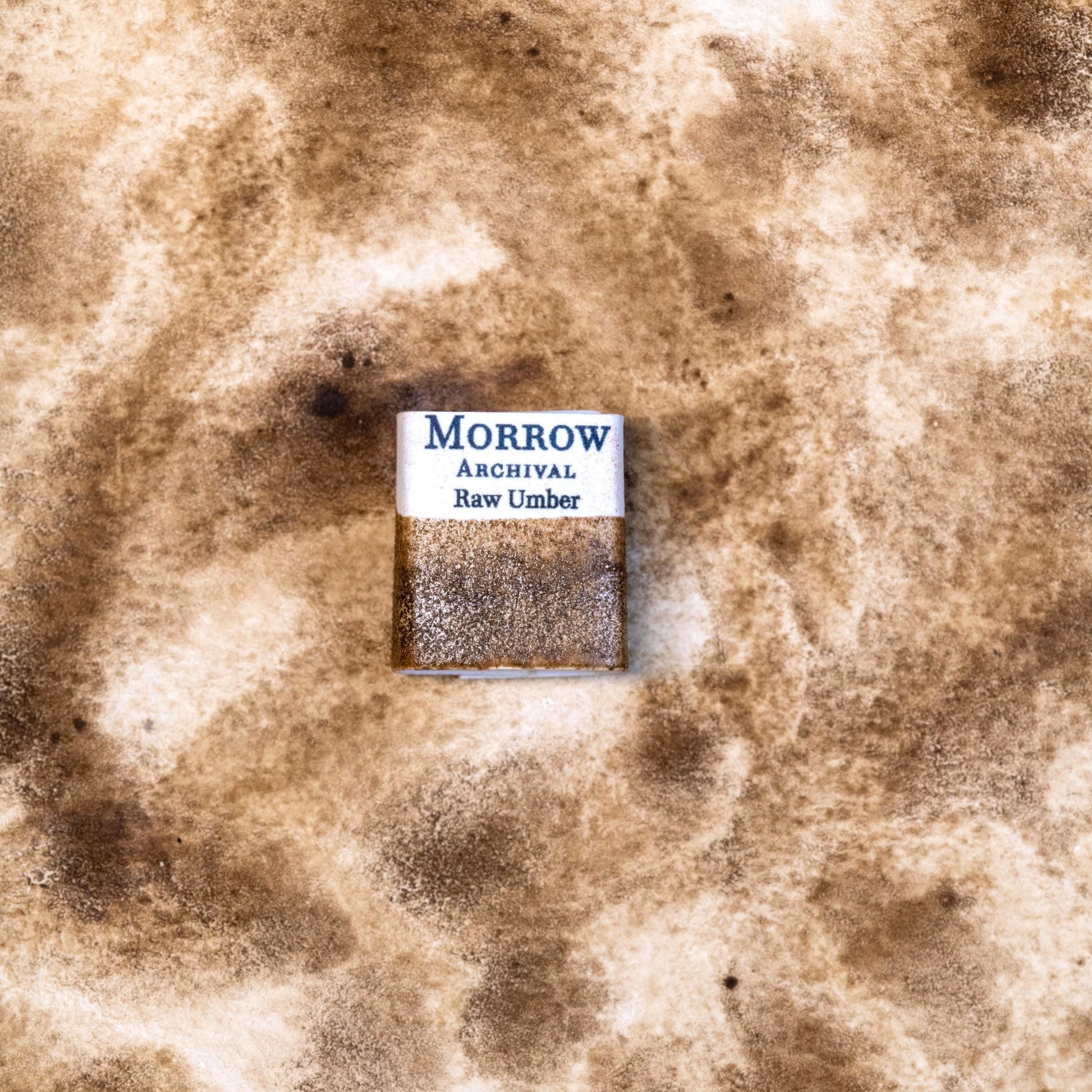 Close-up image of a half pan of Raw Umber handmade watercolor paint, displaying a cool, muted brown tone with a smooth matte finish and gentle granulation. The pan is set on a neutral background, emphasizing the paint‚Äôs natural mineral source and artisanal craftsmanship. Ideal for artists seeking a versatile, earth-based brown for naturalistic and classical work.