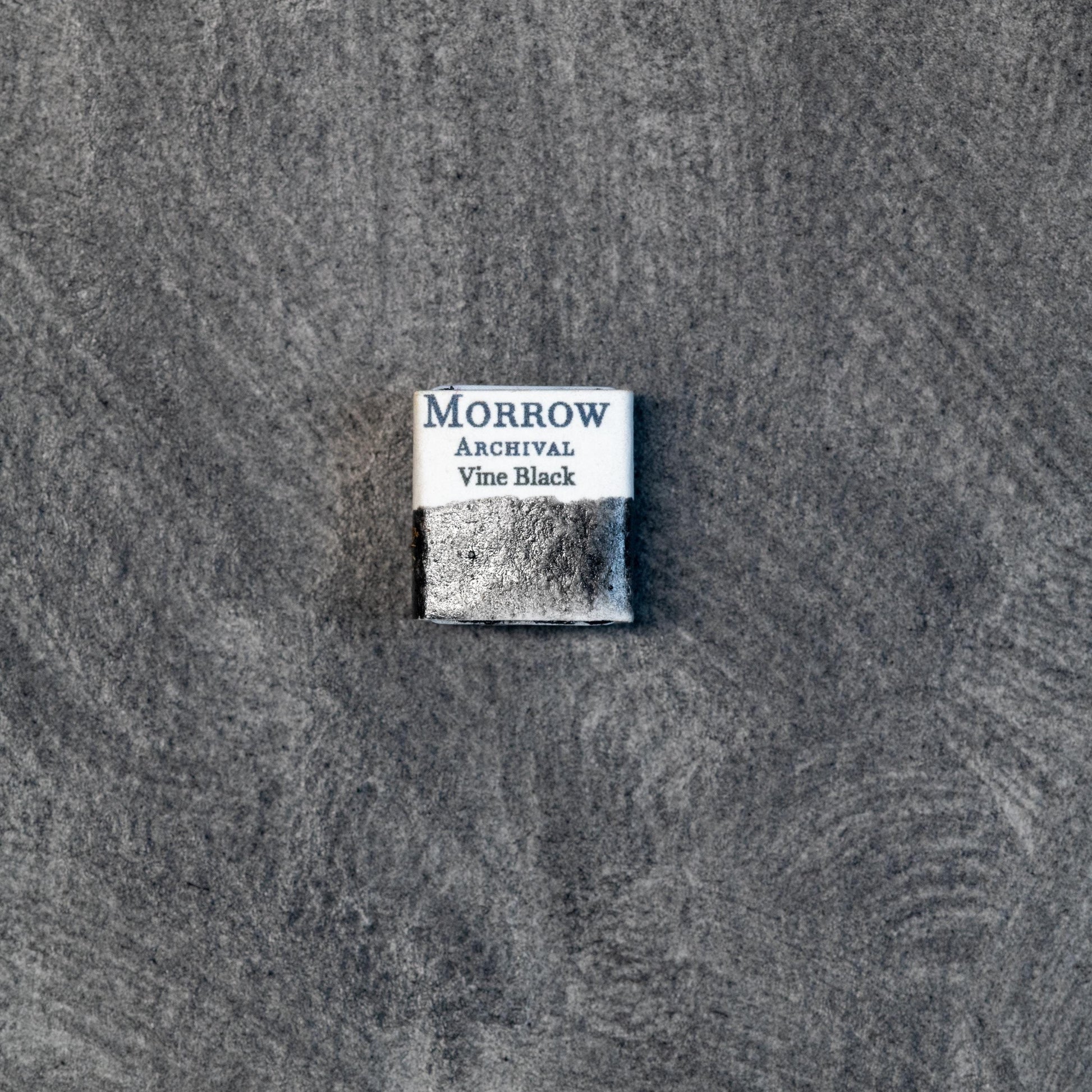 Close-up image of a half pan of Vine Black handmade watercolor paint, showing a soft, charcoal-black hue with a slightly bluish undertone and smooth, semi-transparent surface. The pan rests on a neutral background, emphasizing its natural vegetal origin and artisanal craftsmanship. Ideal for artists seeking a subtle, expressive black for tonal work and historical techniques.