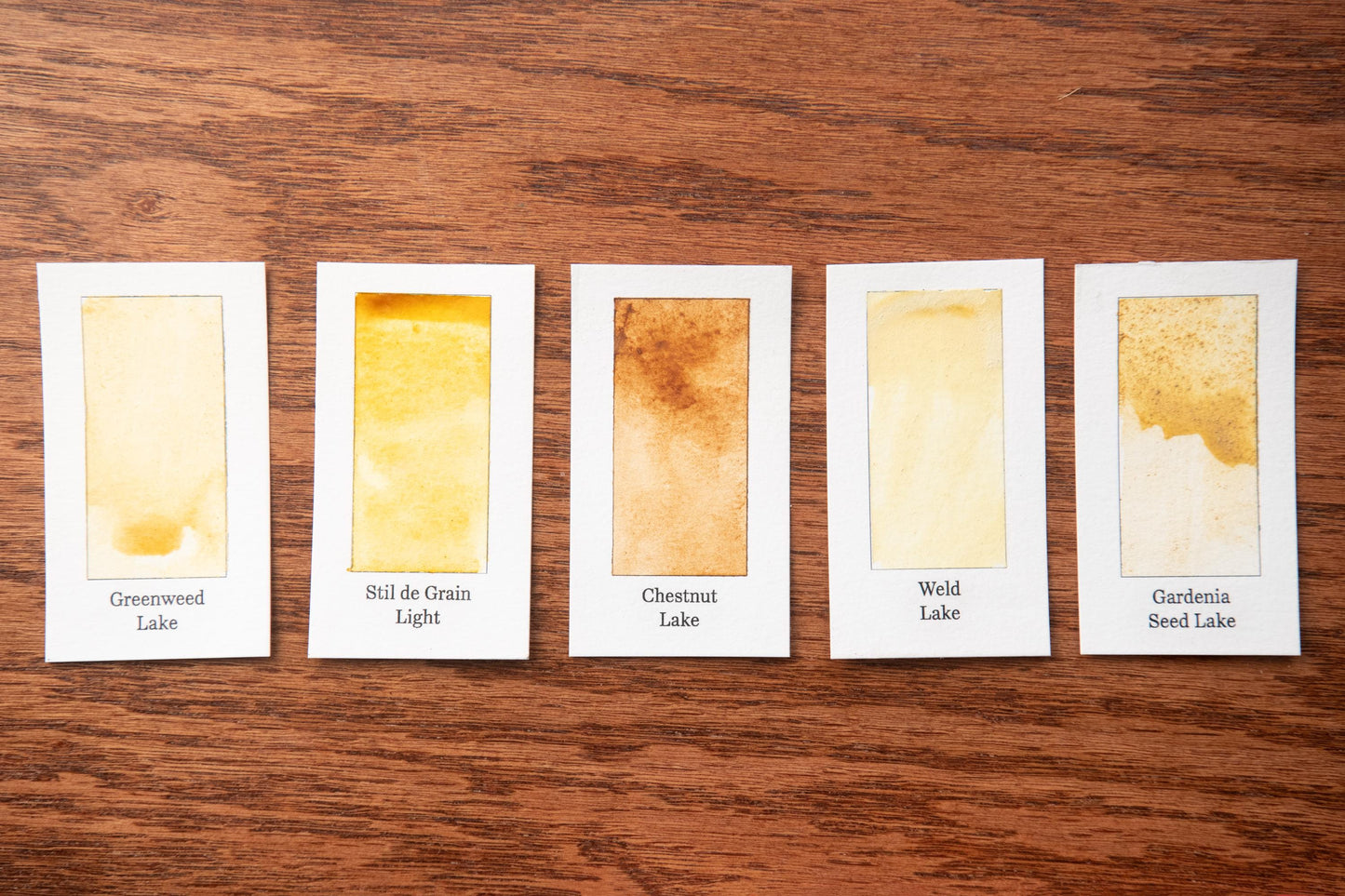 five watercolor paintings on a wooden surface, each with a different color and texture, arranged in a row.