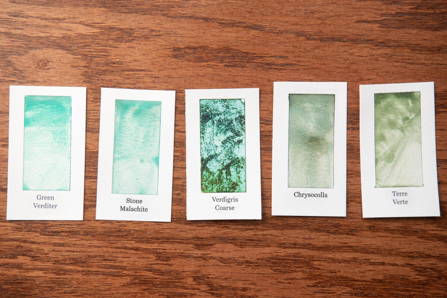 five white cards with different shades of green paint, each labeled with a different type of green paint.