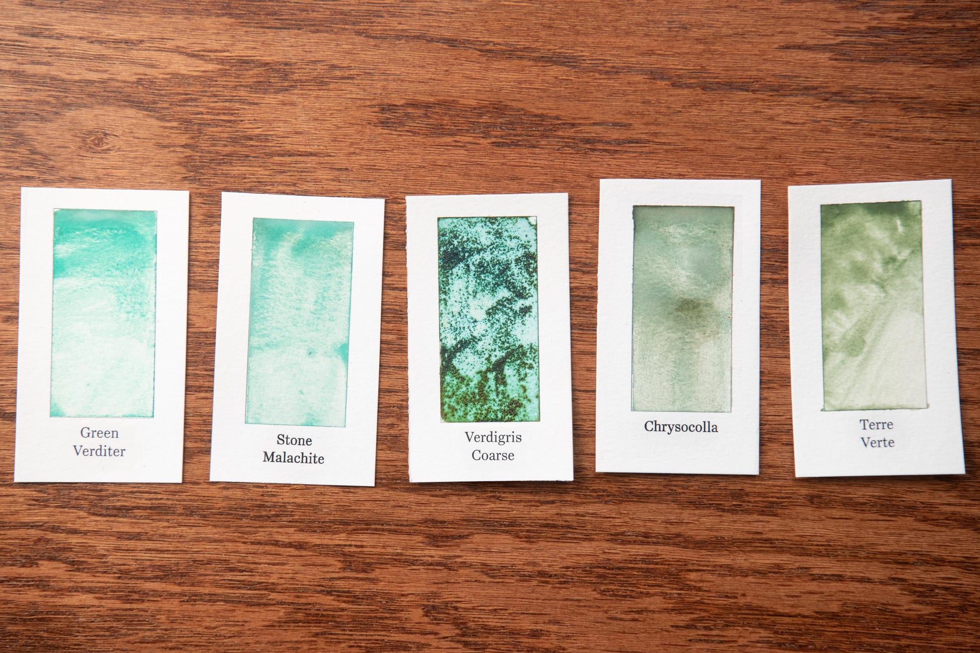 five white cards with different shades of green paint, each labeled with a different type of green paint.