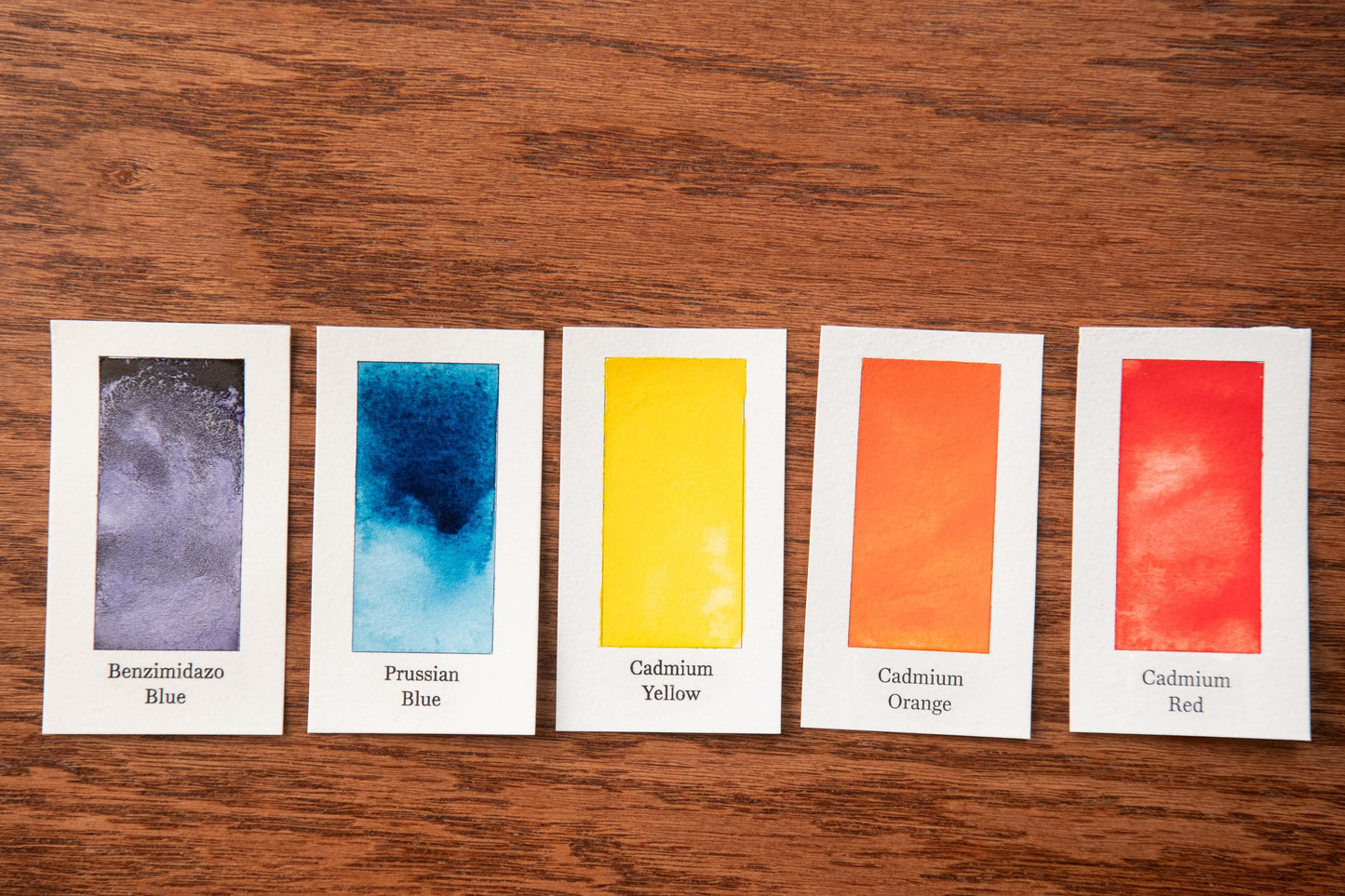 five color swatches arranged on a wooden surface, each representing a different color: blue, yellow, orange, red, and pink.