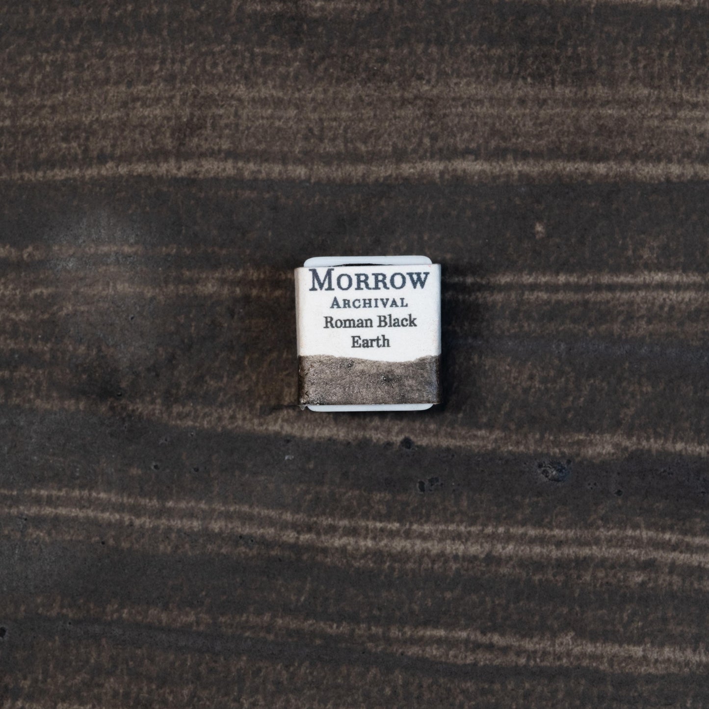 Close-up image of a half pan of Roman Black Earth handmade watercolor paint, displaying a rich, matte black with a subtle earthy texture and natural granulation. The pan is set against a neutral or rustic background, emphasizing its artisanal craftsmanship and natural mineral origins. Ideal for artists seeking a historic, organic black with soft warmth and depth.