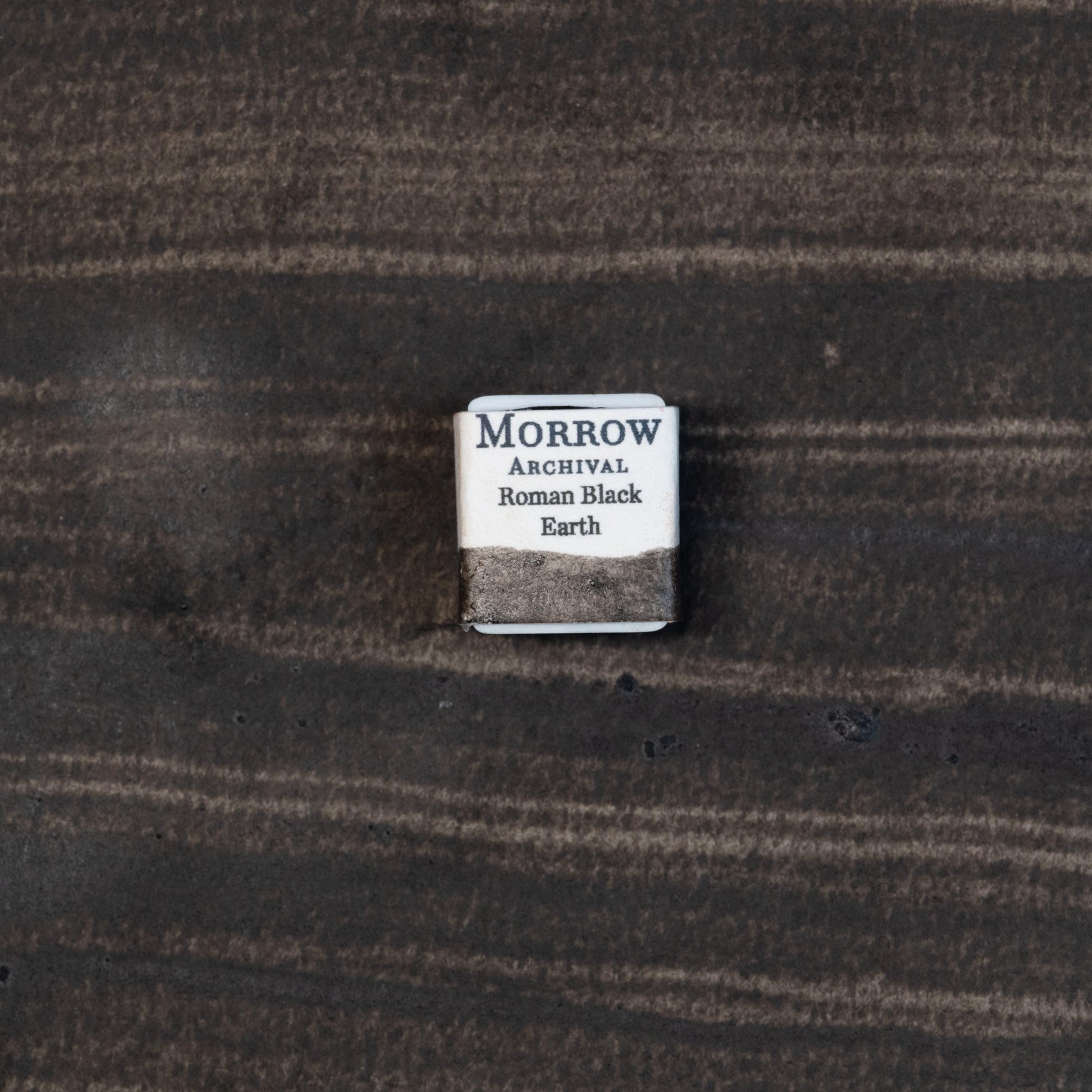 Close-up image of a half pan of Roman Black Earth handmade watercolor paint, displaying a rich, matte black with a subtle earthy texture and natural granulation. The pan is set against a neutral or rustic background, emphasizing its artisanal craftsmanship and natural mineral origins. Ideal for artists seeking a historic, organic black with soft warmth and depth.