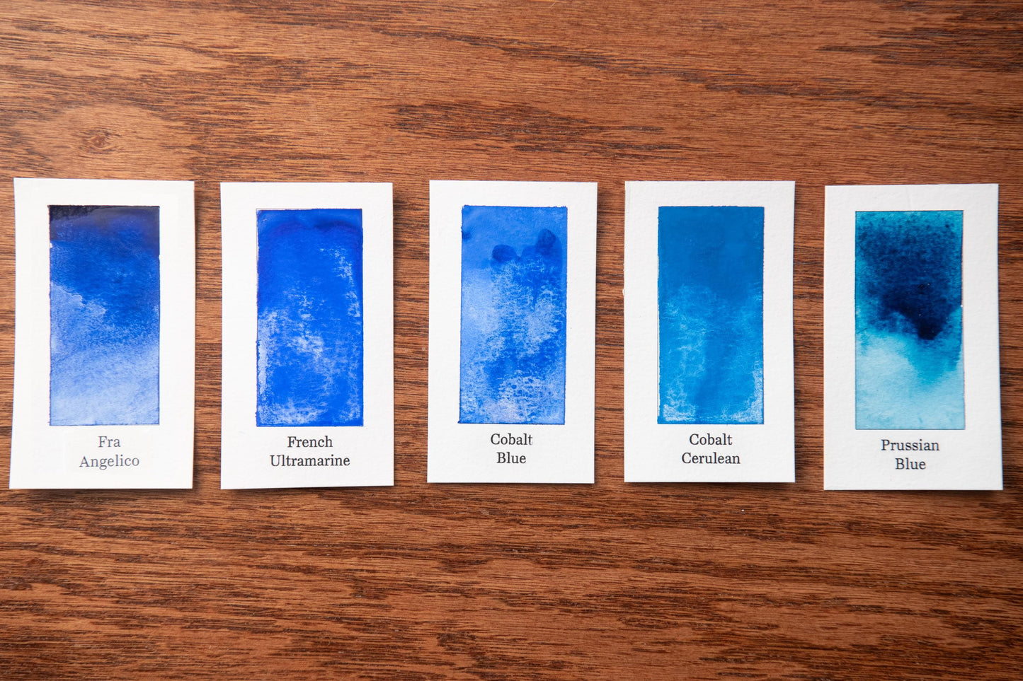 five watercolor paintings in various shades of blue, arranged on a wooden surface.