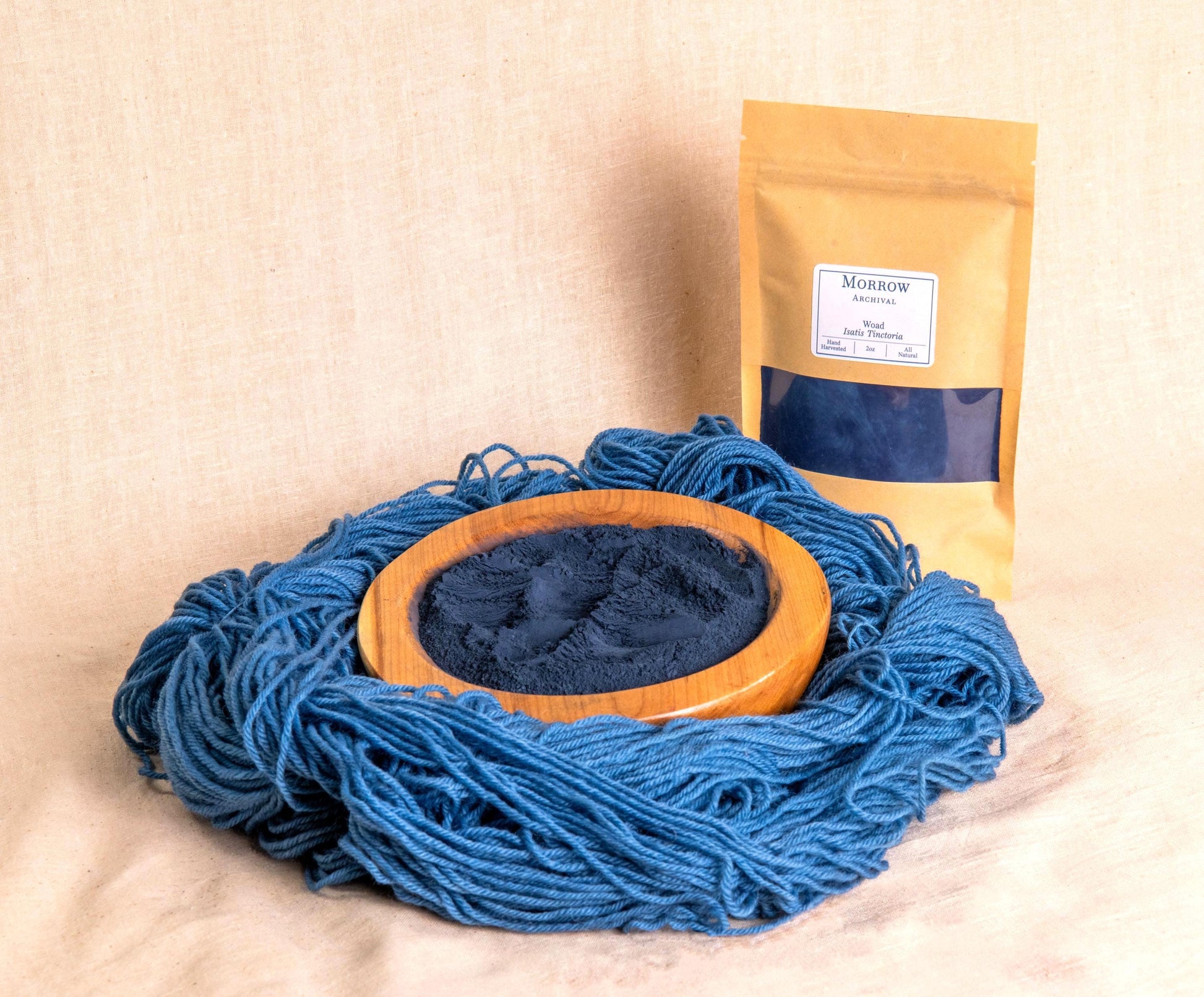 Woad natural dye by Morrow Archival, displayed as a soft blue powder in a wooden bowl, with beautifully dyed blue wool yarn and a labeled kraft pouch in the background. Derived from Isatis tinctoria leaves, this ancient indigo-producing plant has been used since antiquity for natural blue dyes in textiles. Perfect for traditional dyeing, historical reenactment, and eco-conscious fiber artists seeking sustainable, all-natural color.