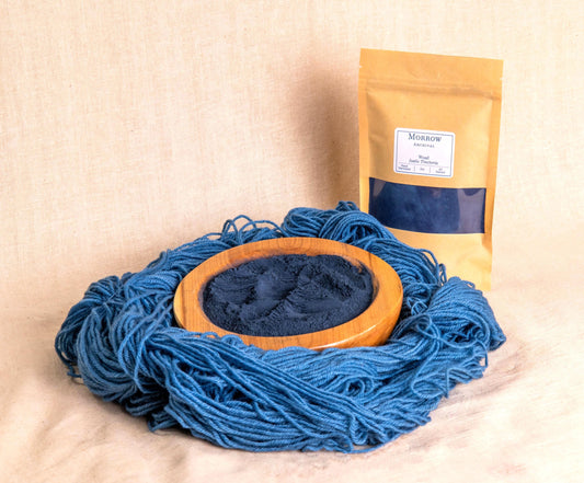 Woad natural dye by Morrow Archival, displayed as a soft blue powder in a wooden bowl, with beautifully dyed blue wool yarn and a labeled kraft pouch in the background. Derived from Isatis tinctoria leaves, this ancient indigo-producing plant has been used since antiquity for natural blue dyes in textiles. Perfect for traditional dyeing, historical reenactment, and eco-conscious fiber artists seeking sustainable, all-natural color.