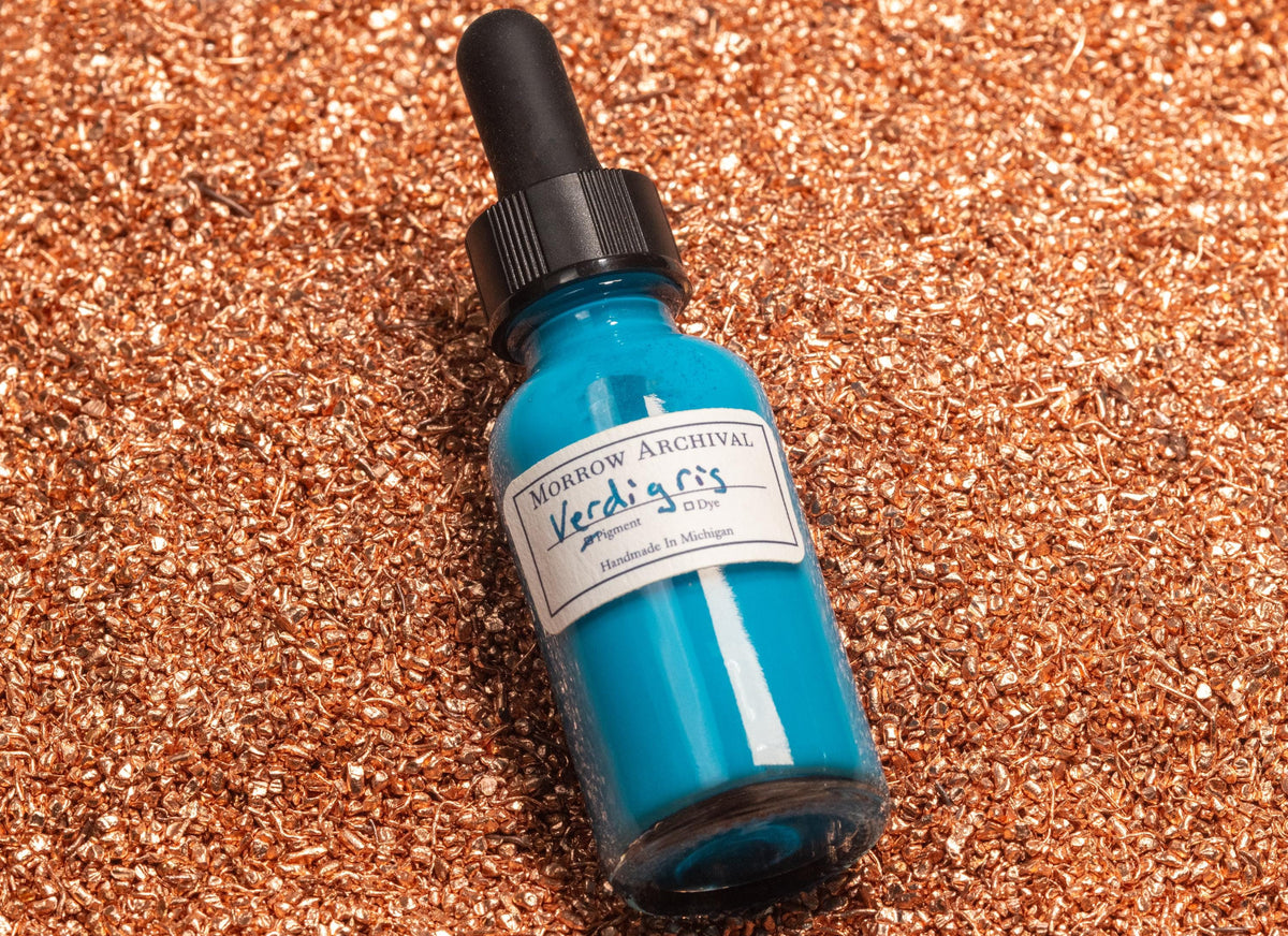 Verdigris ink bottle resting on oxidized copper sheets and fine copper acetate powder, visually tying the vibrant turquoise ink to its chemical-mineral origins. The scene reflects historic methods of pigment creation from patinated metal.
