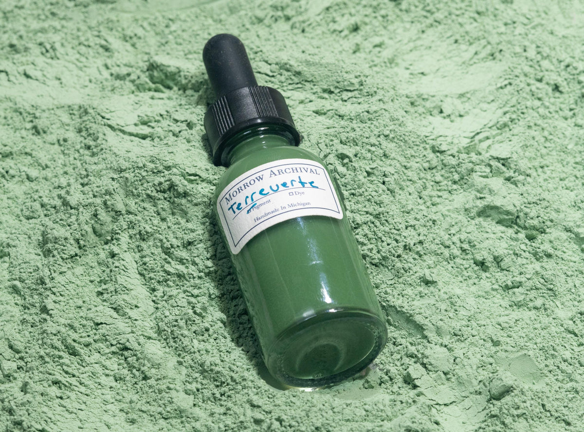 Bottle of Terre Verte ink placed on natural green earth powder and clay nodules. The subdued sage and olive hues of the raw mineral reflect the gentle tone and long artistic history of this pigment in underpaintings and figure work.