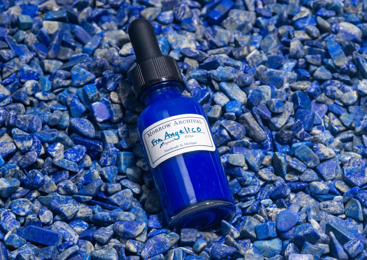 Clear glass bottle of Fra Angelico Blue ink nestled on raw lapis lazuli stones, showing the mineral’s ultramarine blue veins. The iconic semi-precious stone underscores the historic and luxurious origin of this deep celestial pigment.