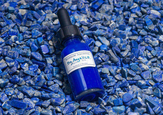 Clear glass bottle of Fra Angelico Blue ink nestled on raw lapis lazuli stones, showing the mineral’s ultramarine blue veins. The iconic semi-precious stone underscores the historic and luxurious origin of this deep celestial pigment.