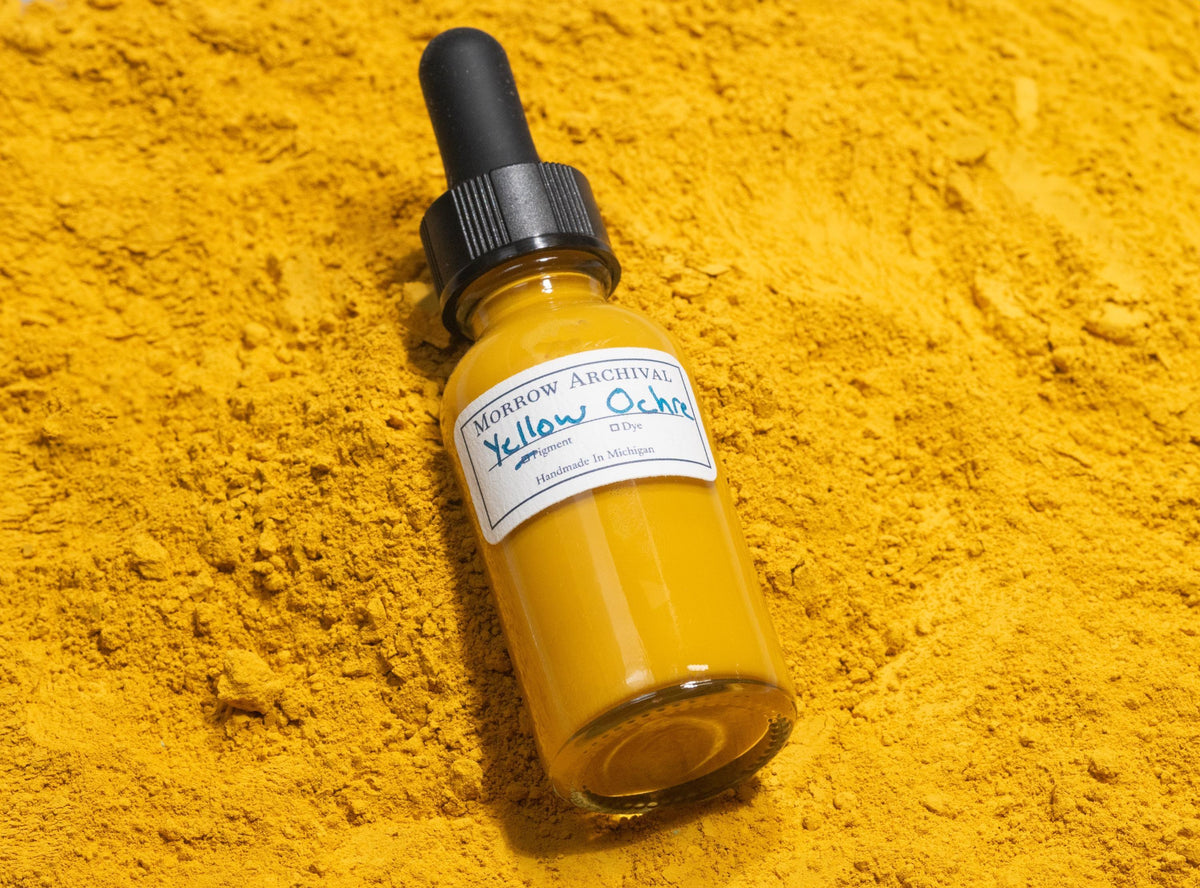 Glass bottle of Yellow Ochre ink placed on yellow ochre stones and powdered pigment. The mineral-rich surface showcases the earthy golden tone’s direct connection to natural iron oxide deposits, a staple pigment in global art history.