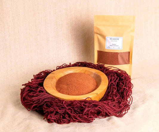 Madder Dye by Morrow Archival, shown as a warm red powder in a wooden bowl, surrounded by vibrant red-dyed wool yarn with a labeled kraft pouch in the background. Made from the roots of Rubia tinctorum, this historic natural dye produces rich reds, pinks, and oranges used since antiquity in textiles from Egypt to India. Perfect for natural dyeing, historical reenactment, and artists seeking eco-friendly, all-natural color.